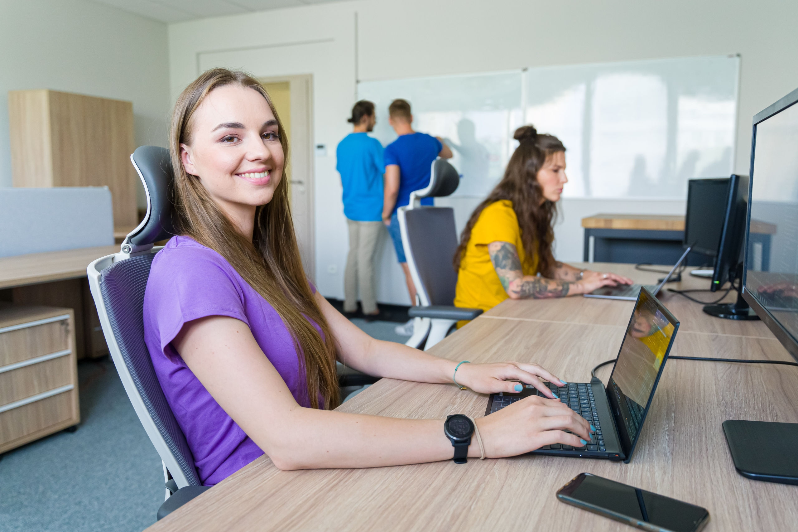A photo of a smiling girl sitting at a desk with a laptop. Behind her, another person is seated at a desk with a laptop, and in the background, two men are standing by a whiteboard