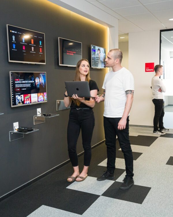 Two people standing and talking to each other in an office corridor. On the wall behind them, four TVs display streaming apps.